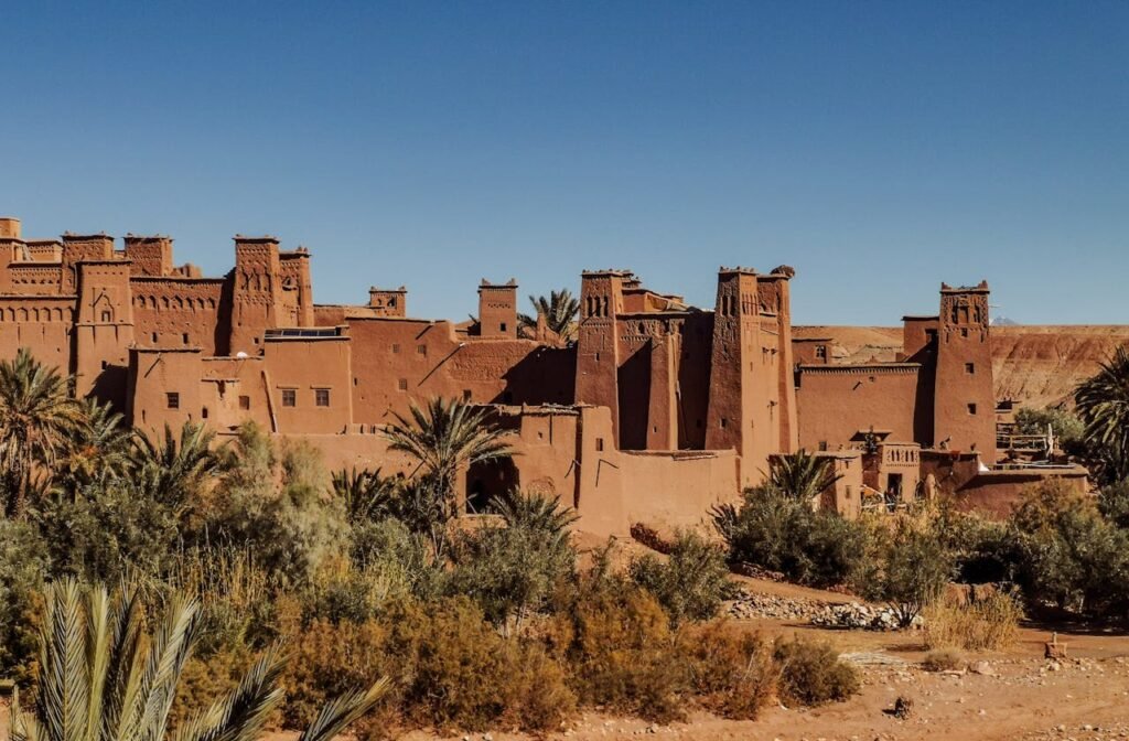 traditons marocain ouarzazat city Exterior of old masonry buildings with square shaped windows near dry sandy terrain with growing palm trees and grass under blue sky